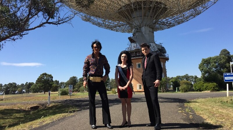 Two Elvis impersonators and a woman standing in front of a big satellite dish in Parkes