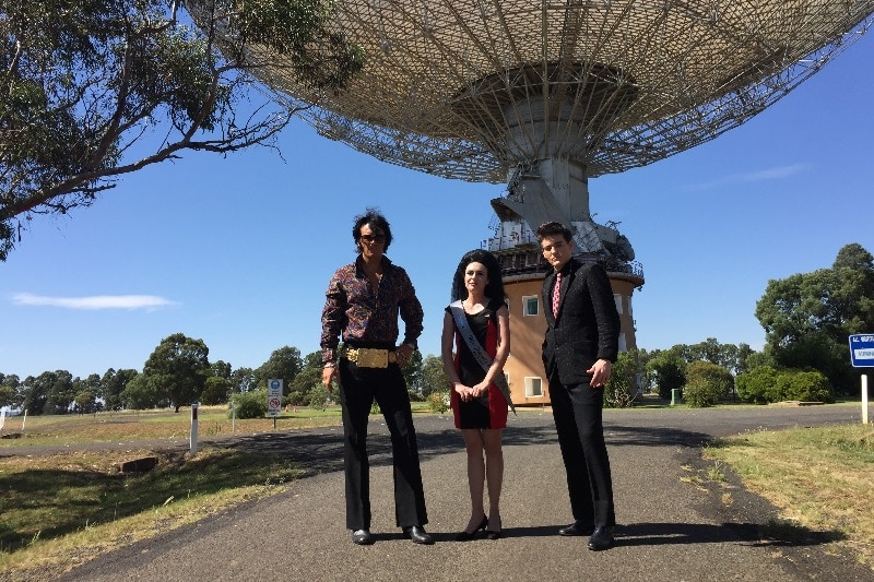Two Elvis impersonators and a woman standing in front of a big satellite dish in Parkes