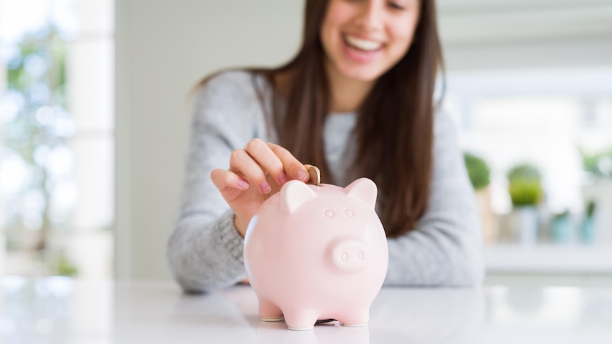 A woman smiles in the background while putting a coin in a pink piggy bank which is in focus.