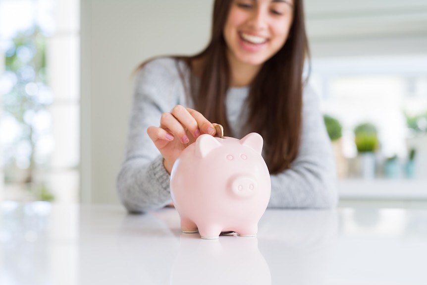 A woman smiles while putting a coin int a pink piggy bank.