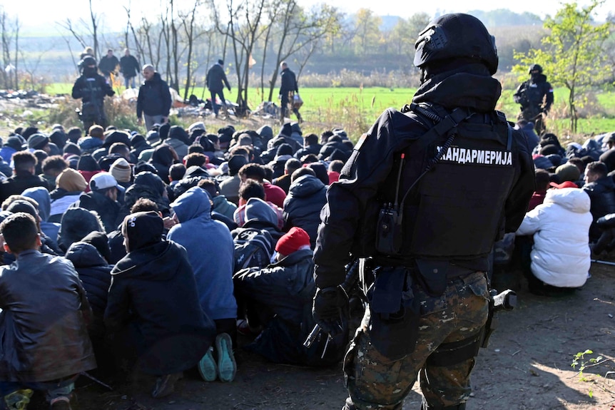 An officer stands in front of a crowd of people crouching on the ground. 