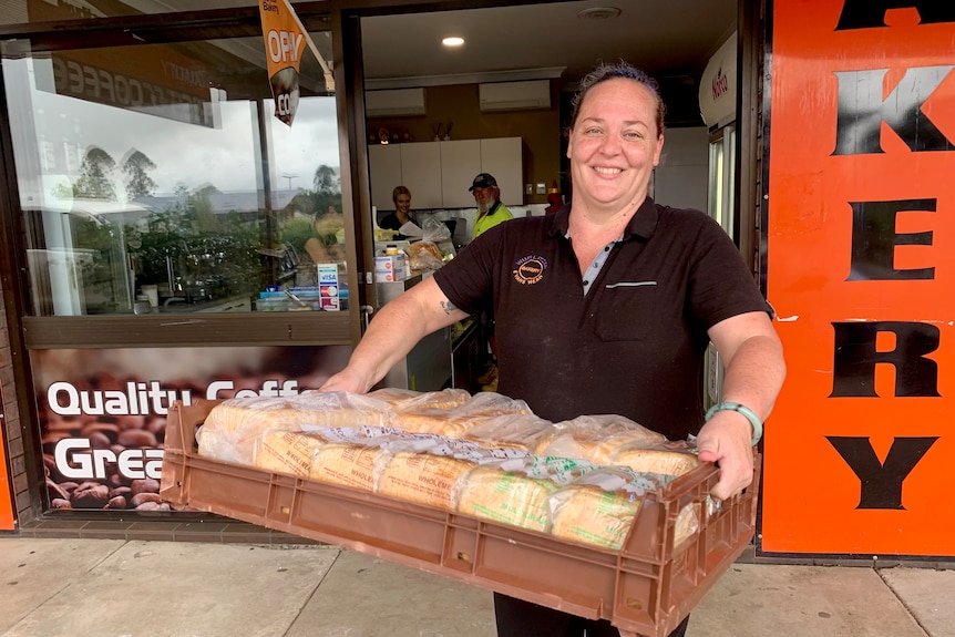 A woman standing outside a bakery holding a tray of fresh loaves of bread.