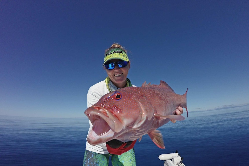 A game fishing operator holds up her impressive catch, a large coral trout, surrounded by blue ocean