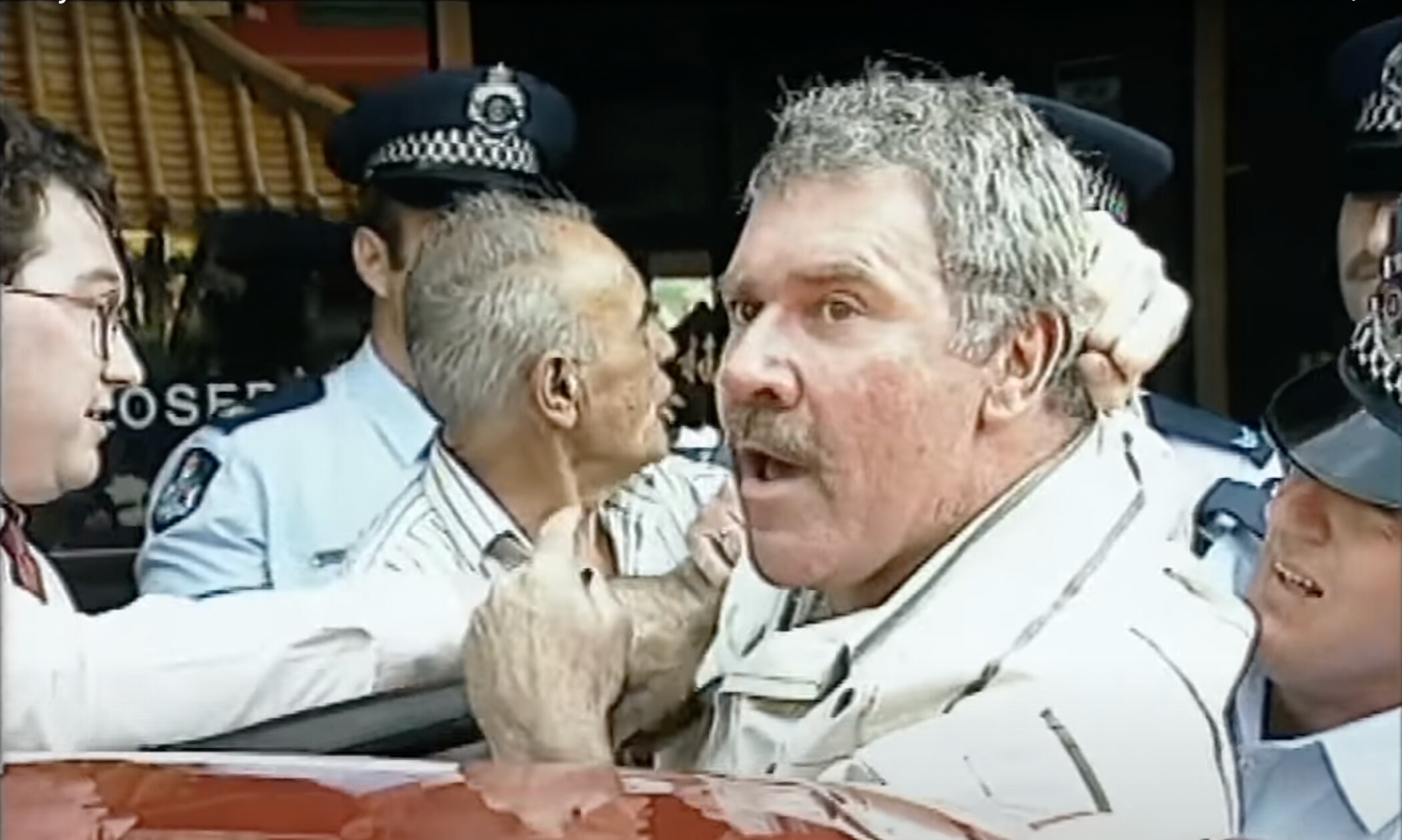 A man looked startrled as he is being grabbed by the hair by a policeman, other policemen surround him.
