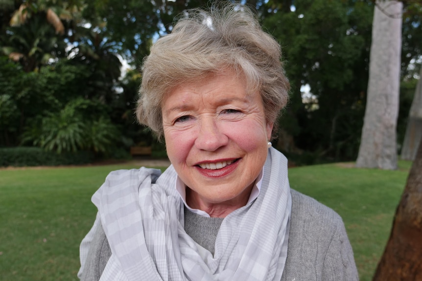 A woman with short blonde hair stands in a green university garden wearing a grey top and scarfe.