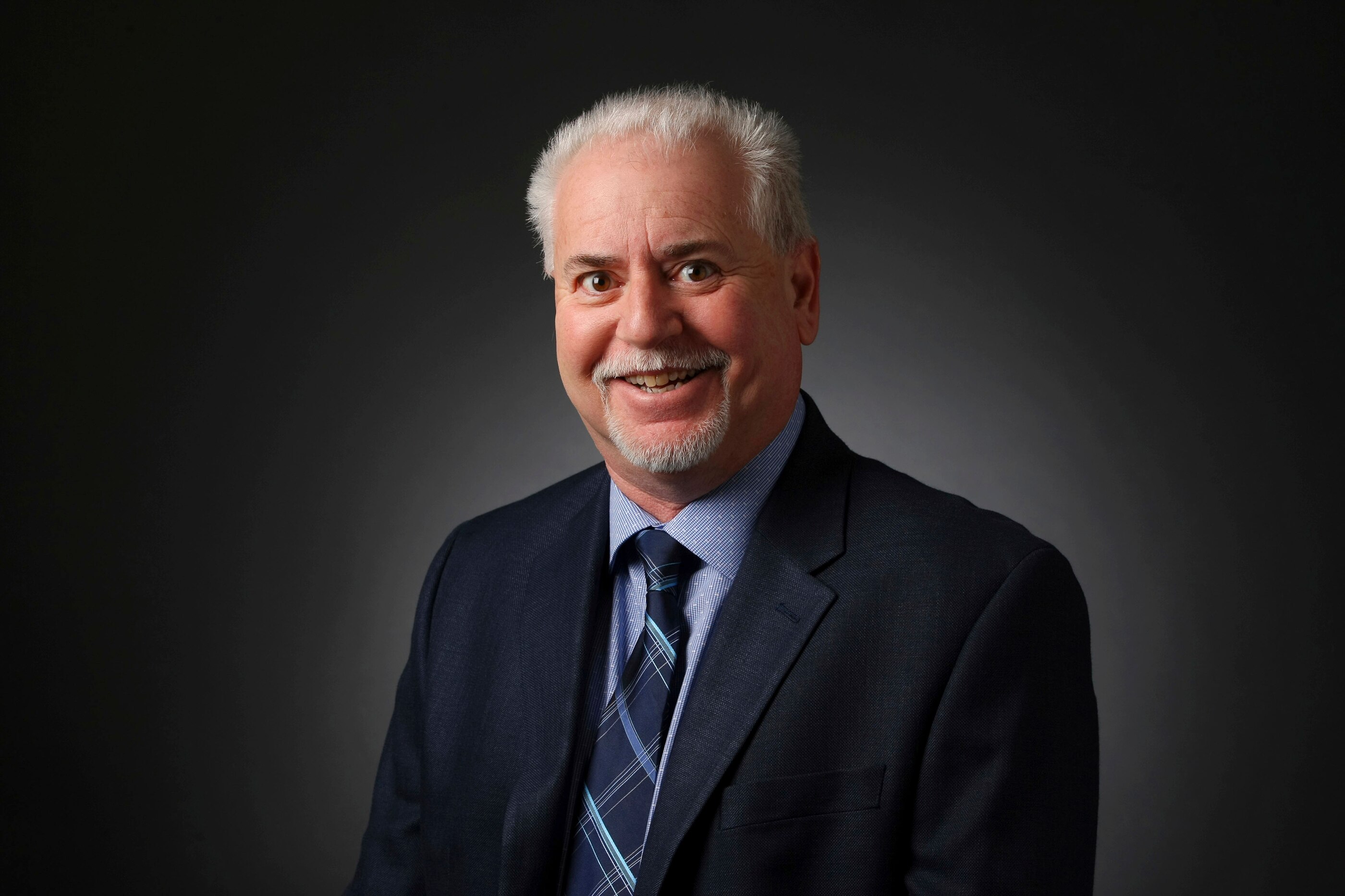A man with grey hair in a suit smiles for a portrait in a studio. 