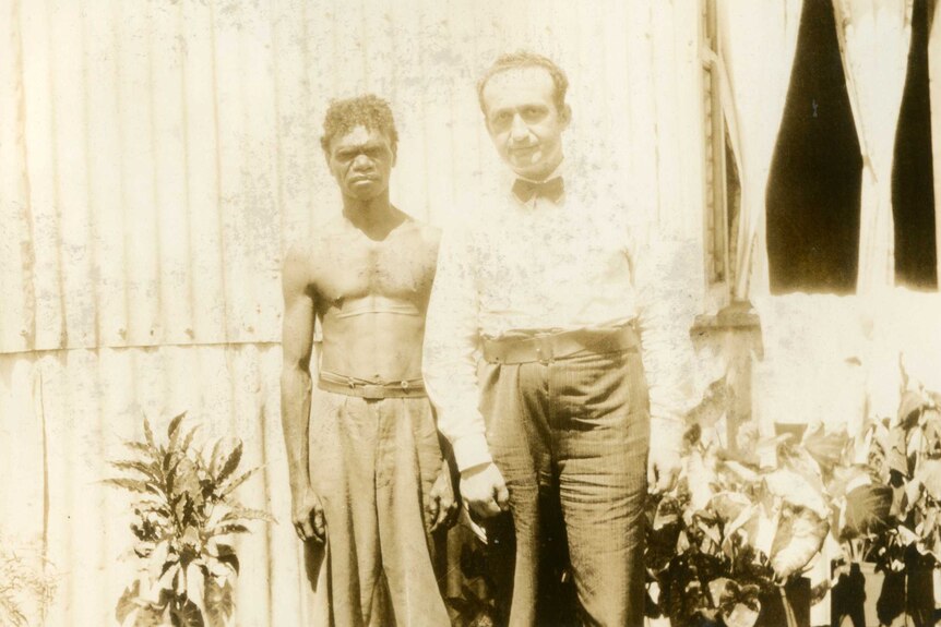 A shirtless aboriginal man who has two long scars on his chest stands next to a white man wearing a bow tie.