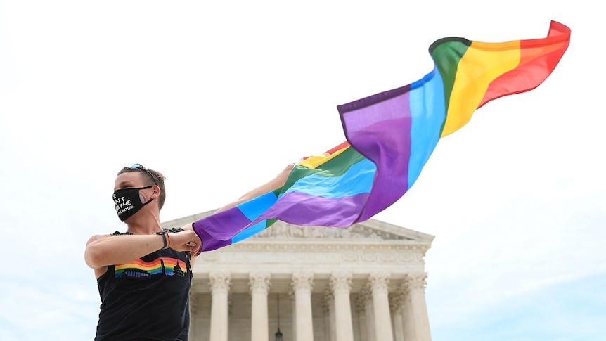 A person wearing a black face mask and singlet holds a Pride Flag in front of a white building.