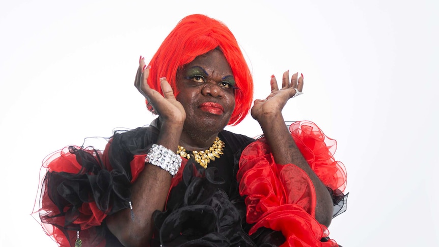 Crystal Love wears a red wig and a red and black dress, and glamourous jewellery. She holds her hands up as she looks up.