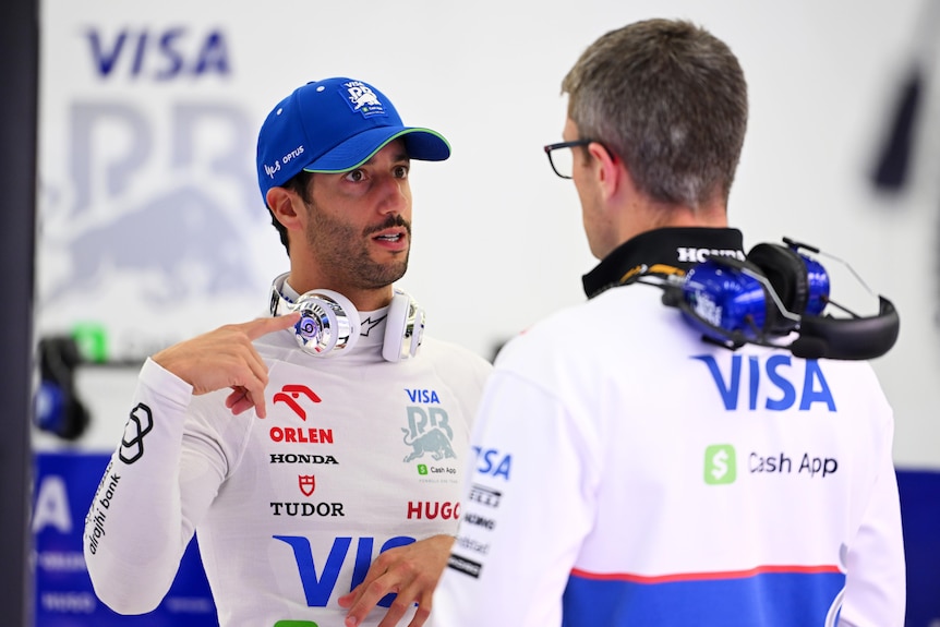 Daniel Ricciardo, in his racing suit, talking to a team member in their F1 garage.