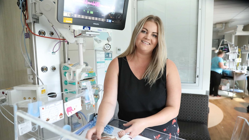 A woman smiles while holding the hand of a premature baby lying in a hospital bed.