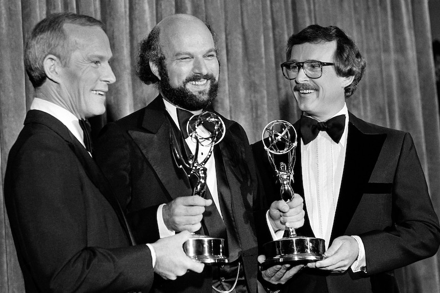 Three men stand holding two Emmys in a black and white image.