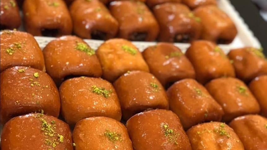 Rows of glazed pastries sprinkled with crumbled pistacchio line a white tray.