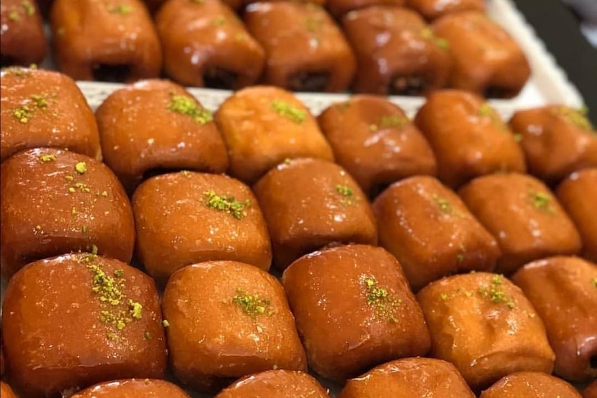 Rows of glazed pastries sprinkled with crumbled pistacchio line a white tray.