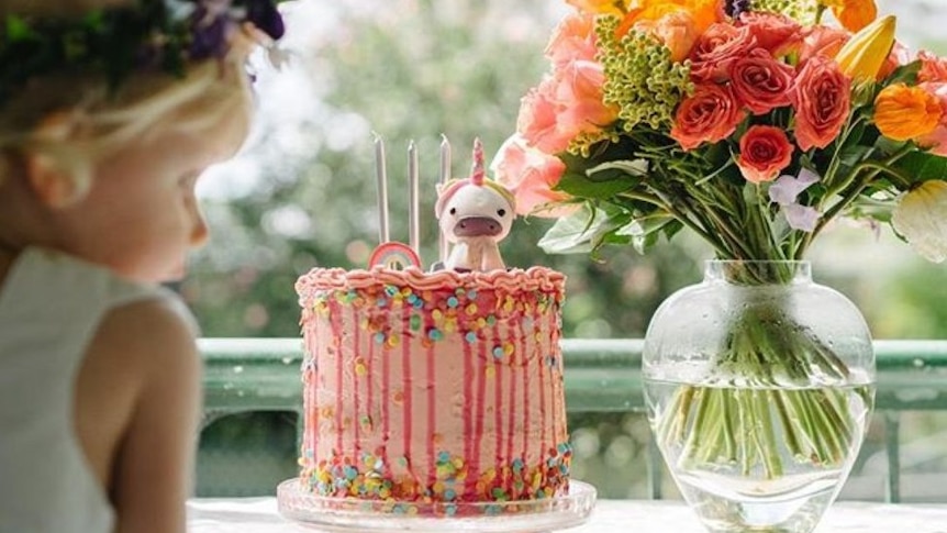 A little girl looks at a birthday cake with a vase of colourful flowers on the table