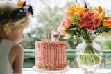 A little girl looks at a birthday cake with a vase of colourful flowers on the table