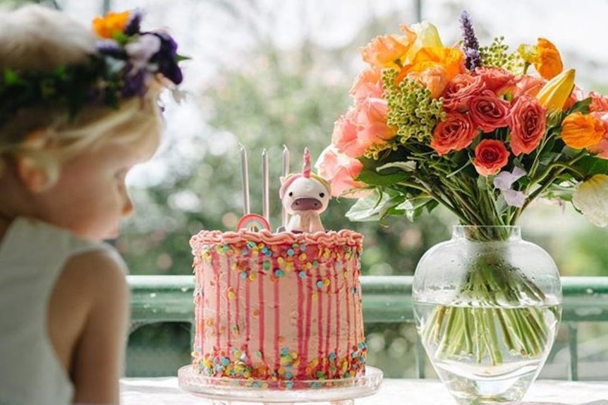 A little girl looks at a birthday cake with a vase of colourful flowers on the table