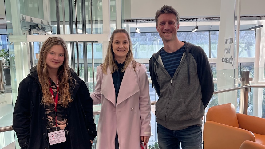 Nahla, Megan and Sammy J stand in the ABC building, in front of large windows, smiling at the camera