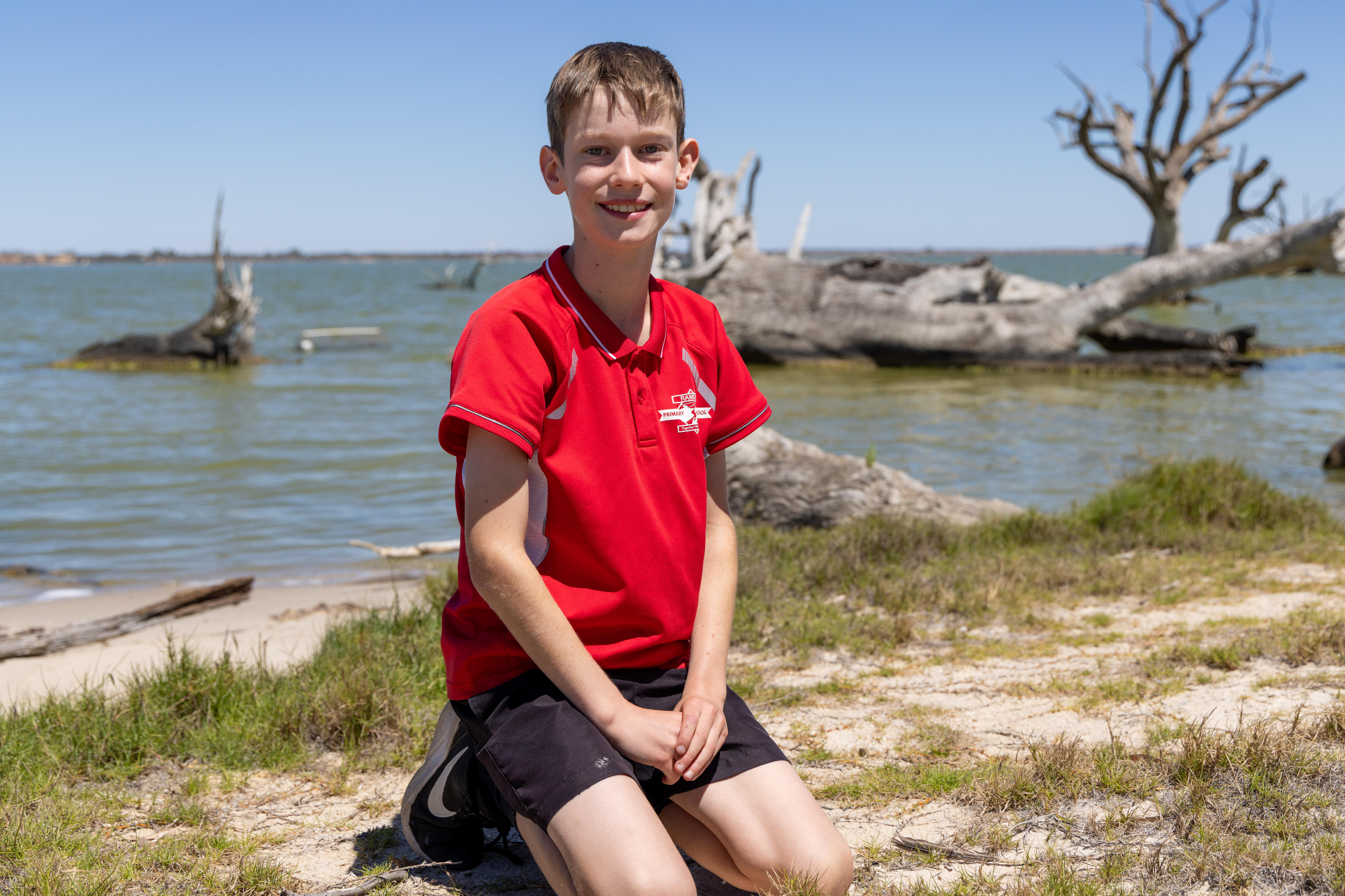 Un estudiante vestido con una camiseta polo roja y pantalones cortos negros, su uniforme escolar, junto a un lago en Riverland.
