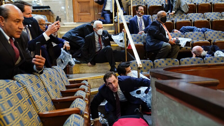 People wearing suits and face masks take cover under chairs while others hold up mobile phones in a seated hall.