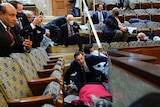 People wearing suits and face masks take cover under chairs while others hold up mobile phones in a seated hall.