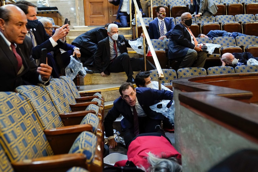 People wearing suits and face masks take cover under chairs while others hold up mobile phones in a seated hall.