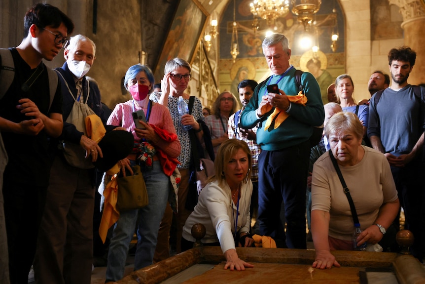Two women knelt down and touching a stone with a group standing behind watching. They are in a church.