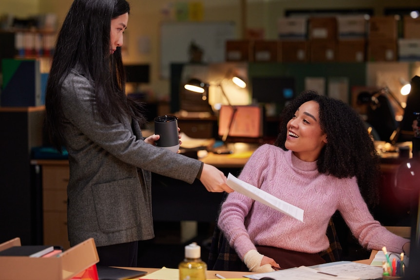 A Chinese Australian woman with long hair and jacket hands a paper to a Black Australian woman sitting a desk in purple jumper