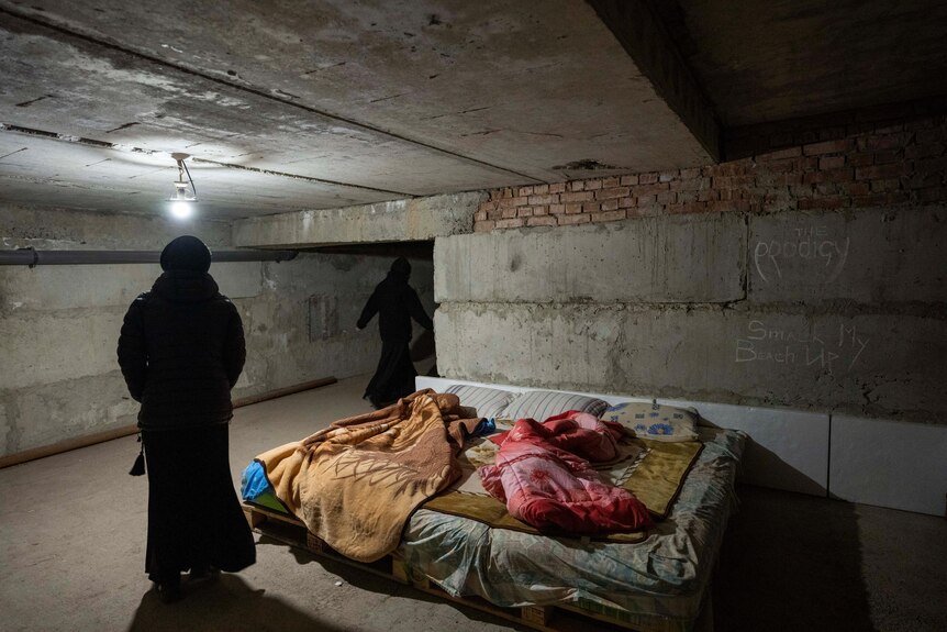 Two nuns walking through a concrete basement, where a bed has been set up.