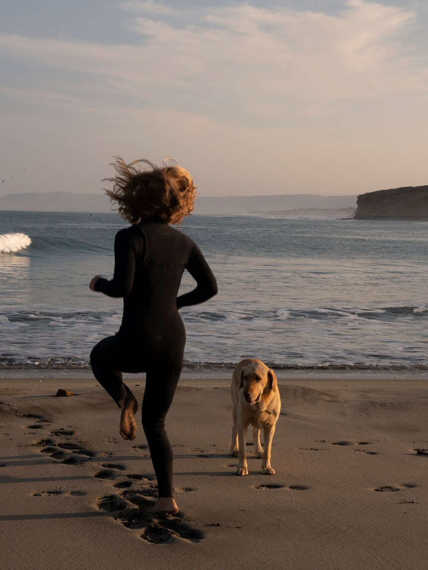 The back of a woman surfer running on the spot on the beach, a dog is next to her.