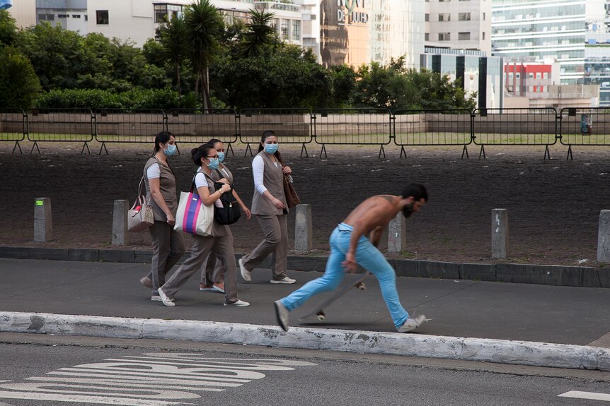 A group of women in face masks walk down a street while a man skateboards past them 