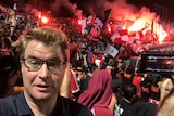 A journalist stands in front of a crowd of soccer fans holding red ad white flags. Flares go off in the distance.