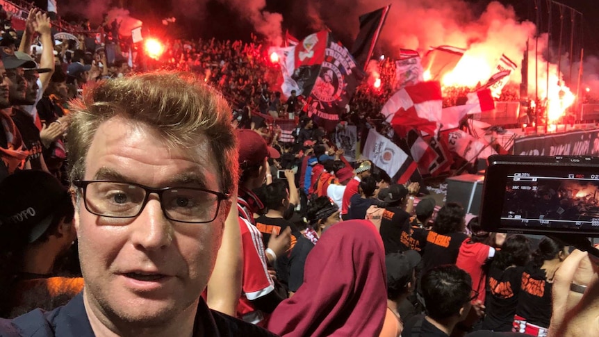 A journalist stands in front of a crowd of soccer fans holding red ad white flags. Flares go off in the distance.