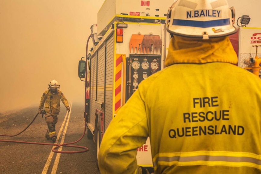 Firefighter carrying a hose in the distance with a truck and another firefighter in foreground.