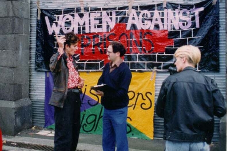 A woman with raised hand speaks to a man taking notes in front of a sign: 'Women against prisons'.