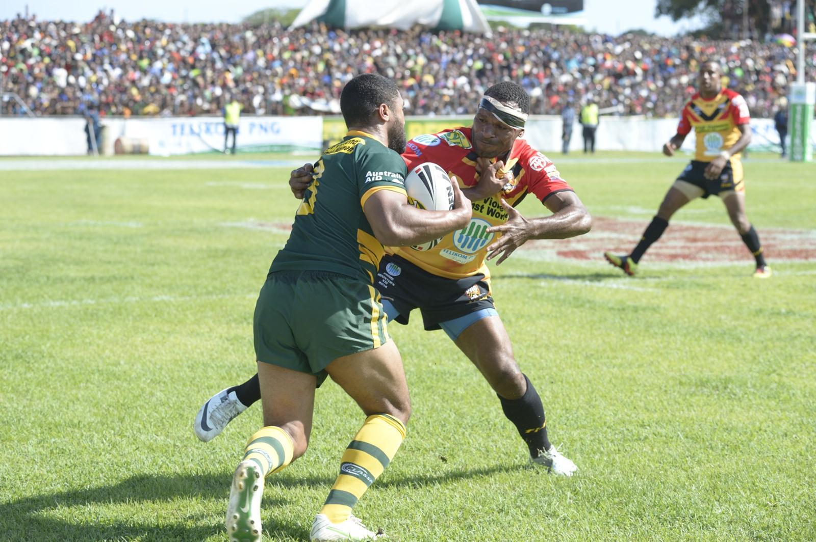 An Australian rugby league player is tackled by a PNG player with a big crowd visible in the background.