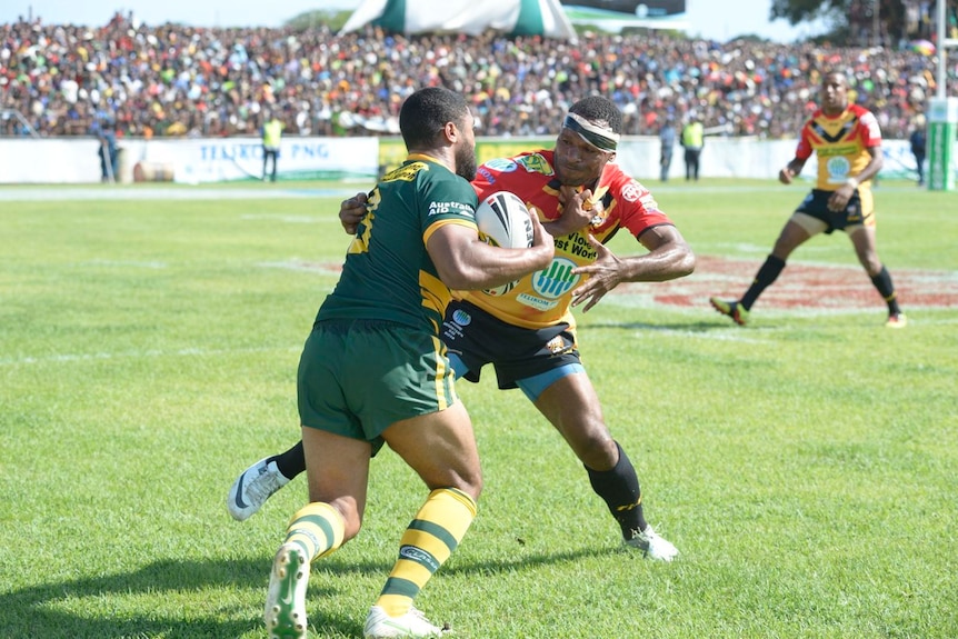 An Australian rugby league player is tackled by a PNG player with a big crowd visible in the background.