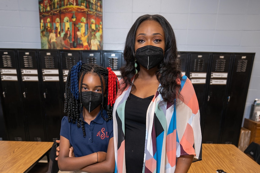 A woman in a black face masks stands next to a girl, also masked, with her arms crossed