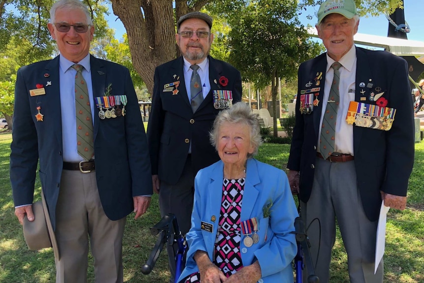 Three elderly men wearing ties, blazers and war medals stand behind an elderly woman, who is also wearing military medals.