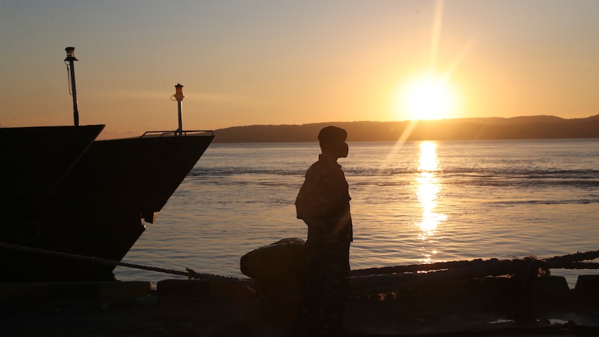 A man standing  by a boat silhouetted by a setting sun