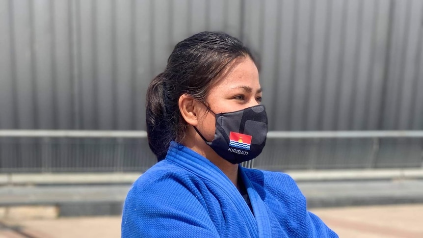 Woman in black mask and blue judo robe looks to right of frame with arms crossed.