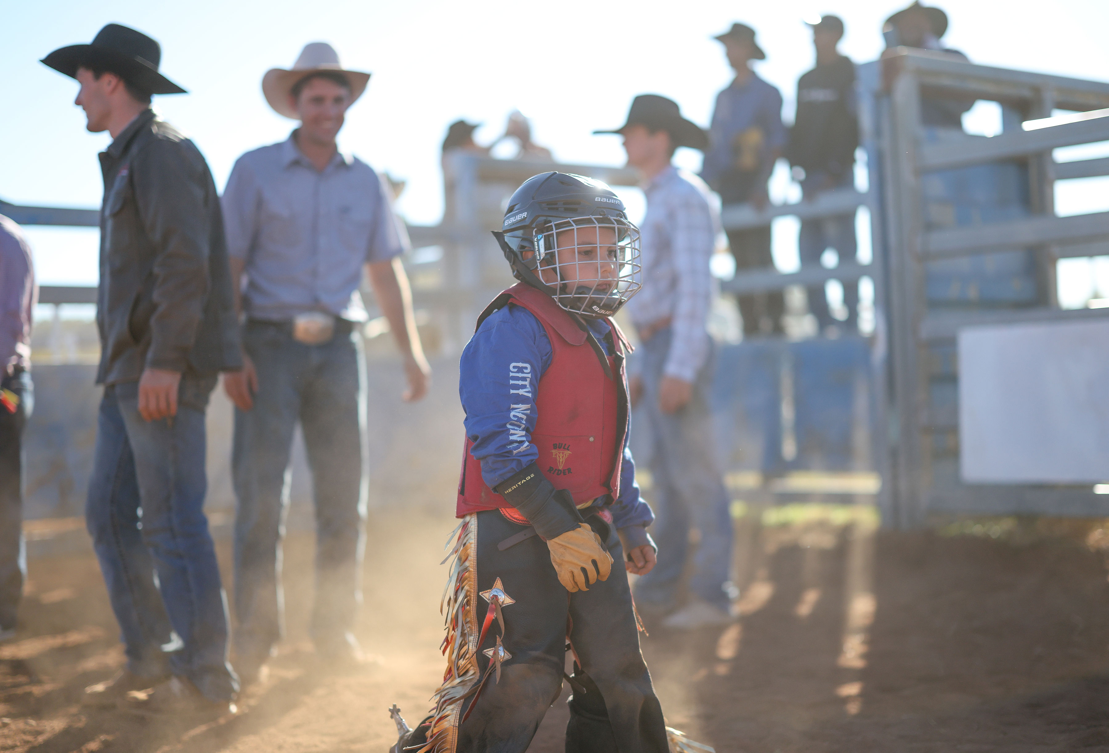 A young rider wearing a helmet and protective vest shakes hands with an adult instructor in a red dirt rodeo arena.