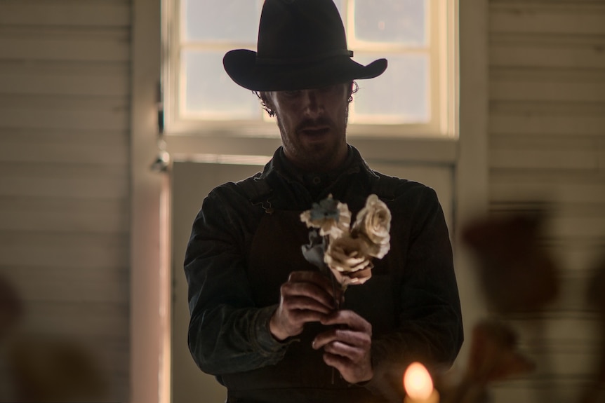 A 40-something man in a cowboy hat stands in shadow holding a bouquet of paper flowers in his hands