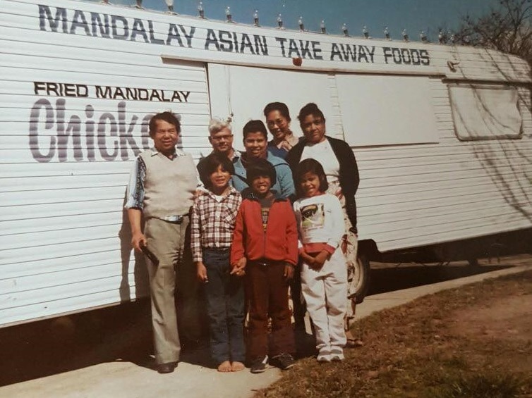 A family stands smiling in front of a white bus selling takeaway Asian food. It is an old photo from decades ago.