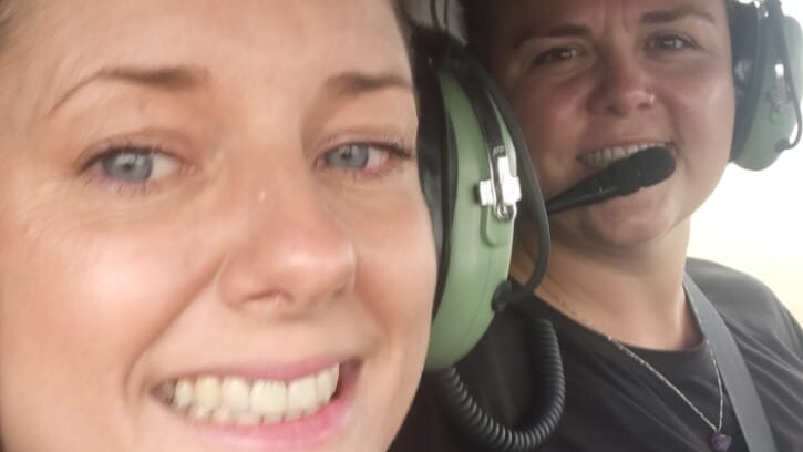 Two women wearing earphones and smiling, strapped into the back of a helicopter.