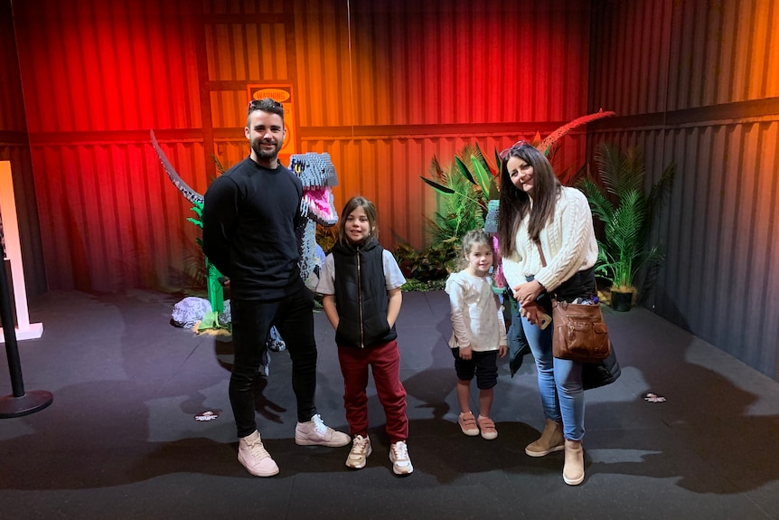 Ben is standing with his two young daughters and mother at an exhibition there in red light and lego dinosaurs in the background