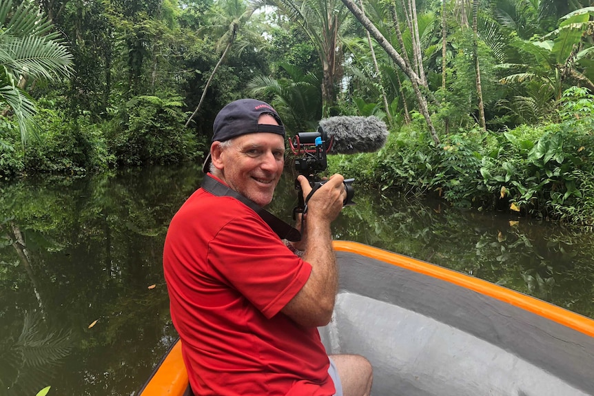 Berkman holding small DSLR camera in a boat on a river surrounded by lush green jungle.