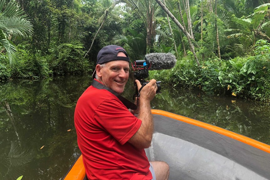 Berkman holding small DSLR camera in a boat on a river surrounded by lush green jungle.