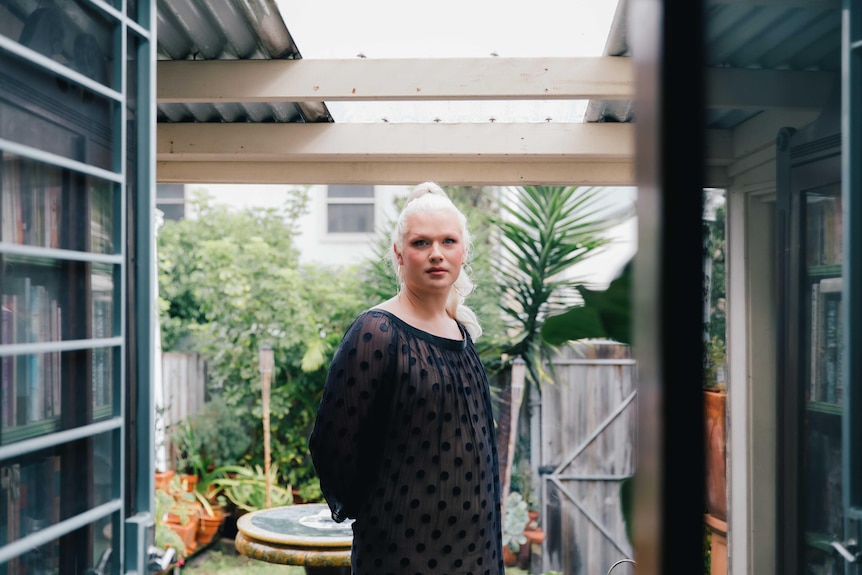 A tall white trans woman with platinum blonde hair stands in a courtyard wearing a sheer black dress.