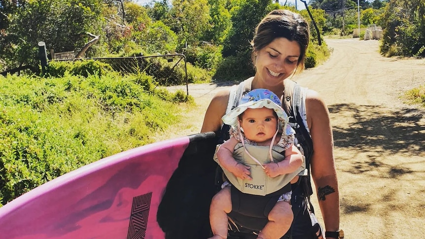 Mez Maconochie carries a baby in a harness while holding a surfboard.